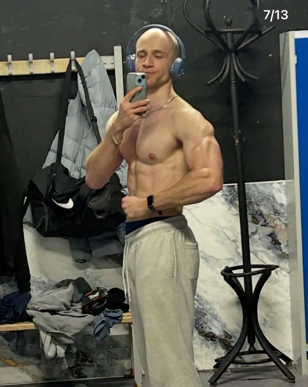 Client in headphones takes a side pose in a gym changing area, wearing grey sweatpants