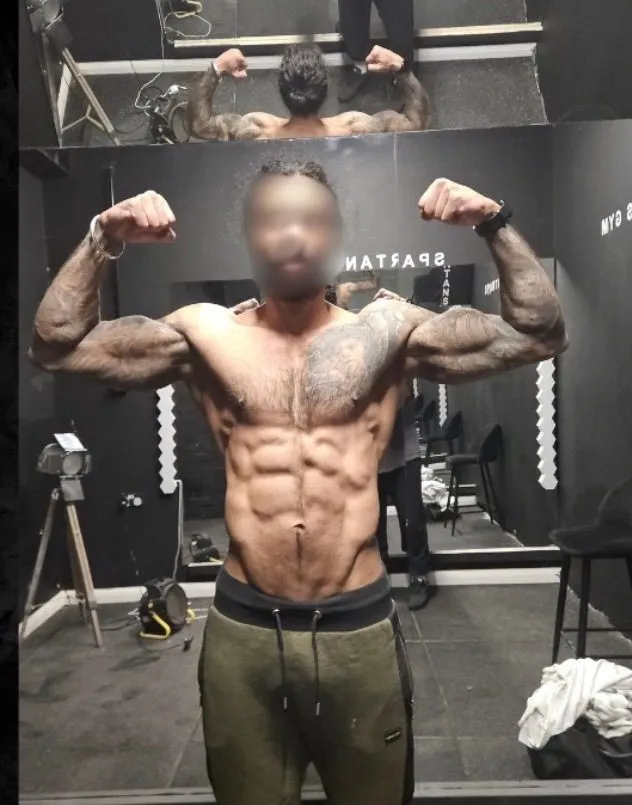Client with a blurred face raises both arms in a front double-biceps pose in a gym mirror, wearing olive joggers