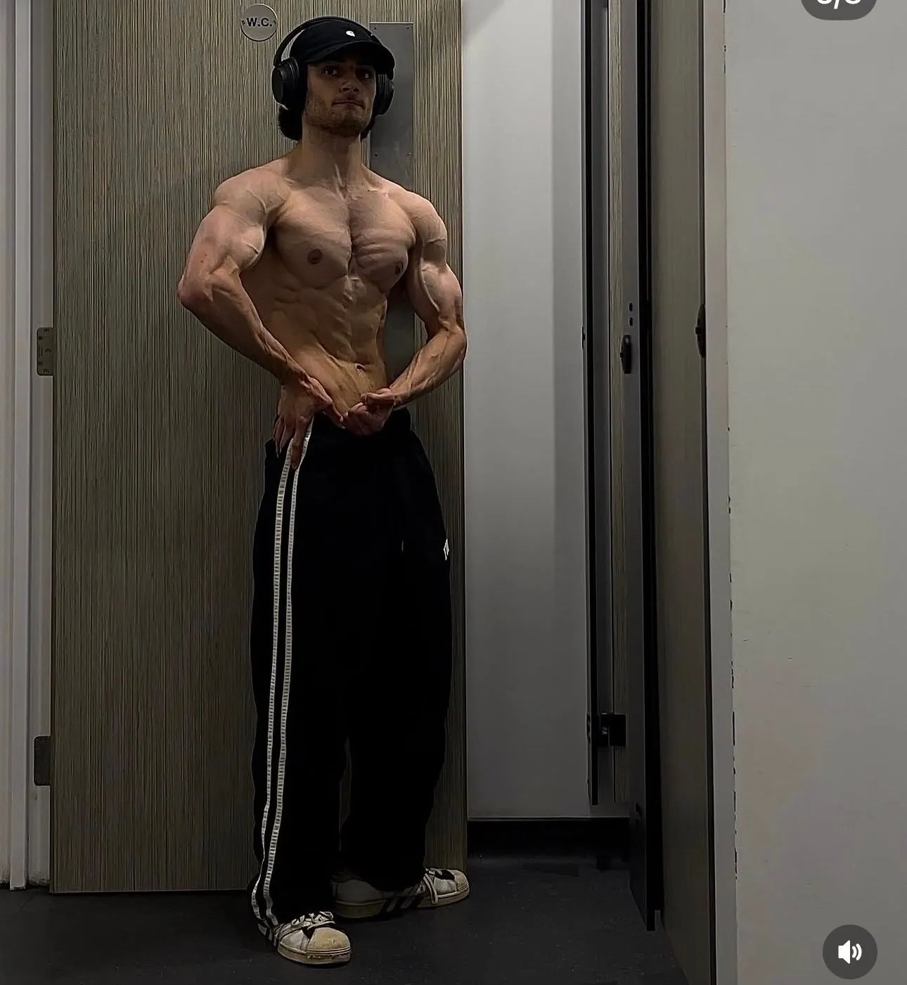 Client wearing headphones and a cap poses in black trousers in a gym changing area, viewed from the side