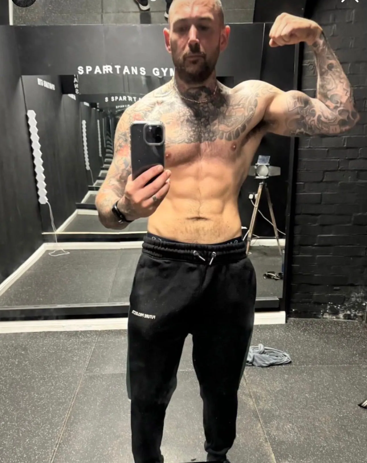 Tattooed client raises one arm in a mirror pose at a gym, wearing black training trousers