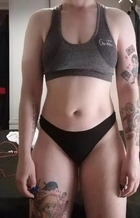 Client stands front-facing in a dark sports bra and black shorts, with tattoos on one arm and thigh